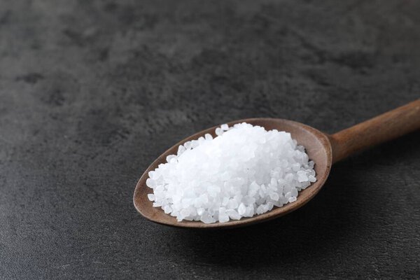 Natural salt in wooden spoon on dark grey table, closeup. Space for text