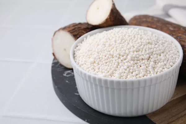 Tapioca pearls in bowl and cassava roots on white tiled table, closeup. Space for text