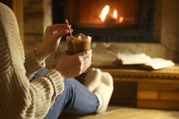 Woman with glass of hot cocoa near fireplace at home, closeup. Space for text