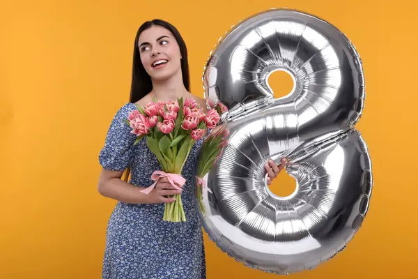 Happy Women\'s Day. Charming lady holding bouquet of beautiful flowers and balloon in shape of number 8 on orange background