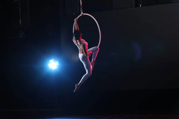 Young woman performing acrobatic element on aerial ring against dark background