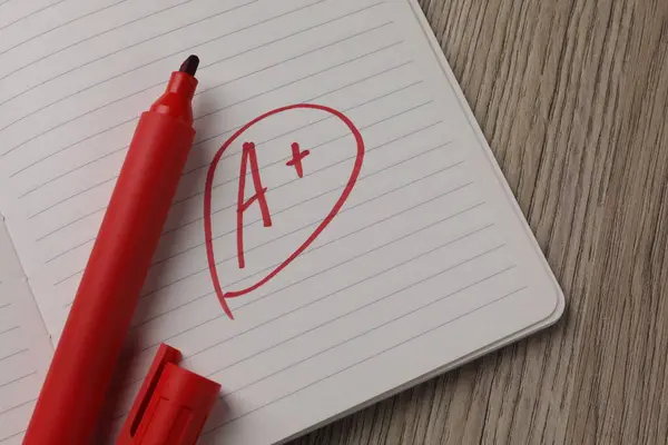 School grade. Red letter A with plus symbol on notebook paper and marker on wooden table, top view