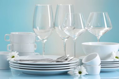 Açık mavi masada bir sürü temiz bulaşık takımı, çatal bıçak, çiçek ve bardak.