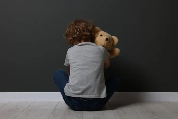 stock image Child abuse. Upset boy with toy sitting on floor near grey wall, back view