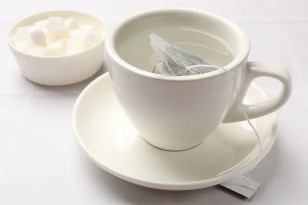 Tea bag in cup with hot water and bowl with sugar on white tiled table, closeup