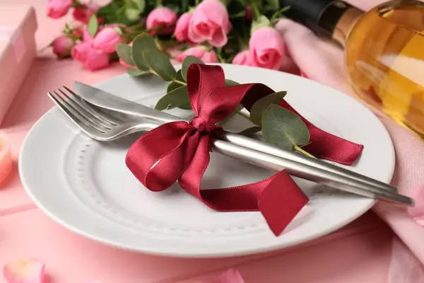 Romantic place setting. Plate, cutlery, eucalyptus branch and roses on pink table, closeup