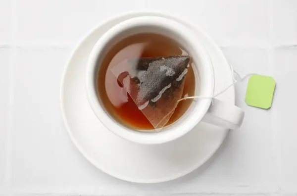 Tea bag in cup with hot drink on white tiled table, top view
