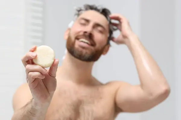 Happy man showing solid shampoo bar in bathroom, selective focus