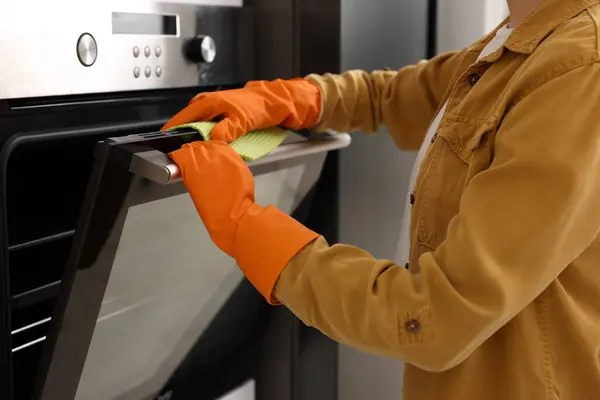 Woman with microfiber cloth cleaning electric oven in kitchen, closeup
