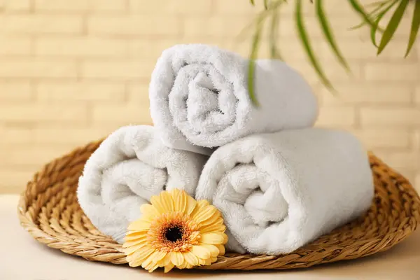 Rolled terry towels, flower and green leaves on white table near brick wall indoors, closeup