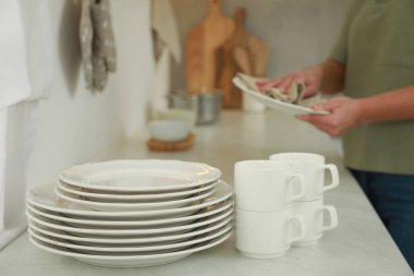 Mutfaktaki beyaz masada tabağı havluyla silen kadın, bulaşık takımına odaklan.