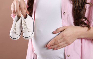 Bej arka planda bebek ayakkabısı tutan hamile kadın, yakın plan.