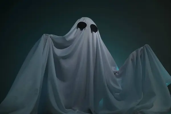 Creepy ghost. Woman covered with sheet on dark teal background, low angle view