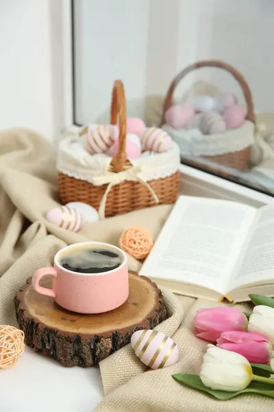 Easter decorations. Cup of coffee, painted eggs, tulips and book on windowsill