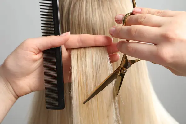 Hairdresser cutting client's hair with scissors on light grey background, closeup