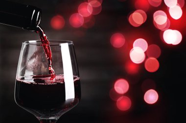 Pouring red wine into glass against dark background with blurred lights, closeup. Space for text clipart