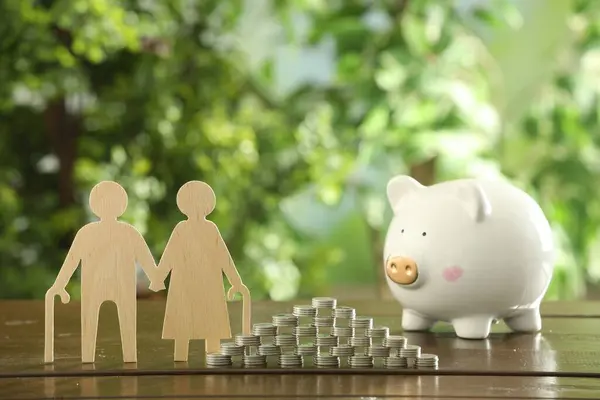 Pension savings. Figure of senior couple, piggy bank and stacked coins on wooden table outdoors
