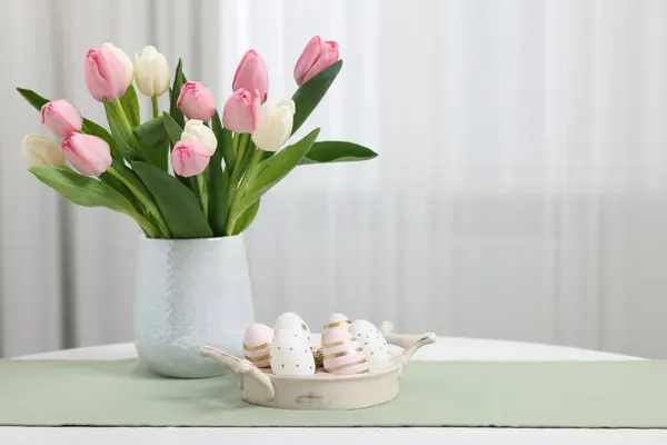 Easter decorations. Bouquet of tulips and painted eggs on white table indoors. Space for text