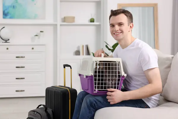 Travel with pet. Man holding carrier with cute cat on sofa at home, space for text