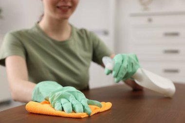 Elinde sprey şişesi ve mikrofiber bez olan bir kadın odasındaki ahşap masayı temizliyor.
