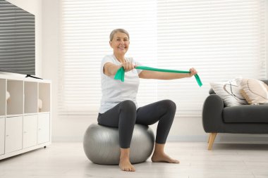 Kıdemli kadın evde spor salonunda elastik direniş bandıyla egzersiz yapıyor.