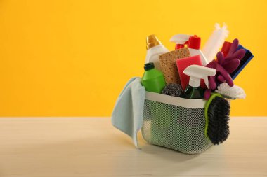 Tahta masadaki sepetteki farklı temizlik ürünleri sarı arkaplan, metin için yer