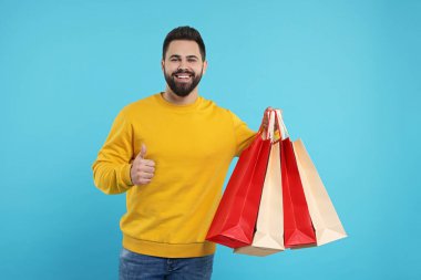 Bir sürü kese kağıdıyla mutlu bir adam açık mavi arka planda baş parmağını gösteriyor.