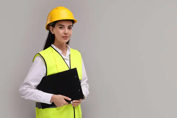 Engineer in hard hat holding clipboard on grey background, space for text