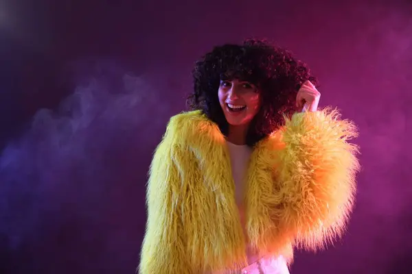 ネオンライトと煙の色の背景に黄色い毛皮のコートの美しい若い女性 — ストック写真