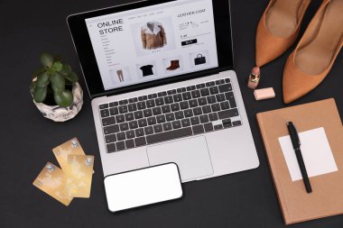 İnternetteki web sitesi laptop ekranında. Bilgisayar, akıllı telefon, kredi kartları, kadın ayakkabıları, kırtasiye malzemeleri ve siyah arkaplanda ruj, üst görünüm