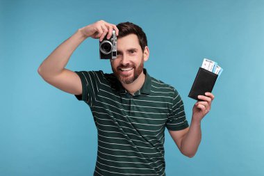 Pasaportu, kamerası ve açık mavi arka planda biletleri olan gülümseyen adam.