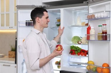 Mutfakta buzdolabının yanında elma tutan mutlu adam.