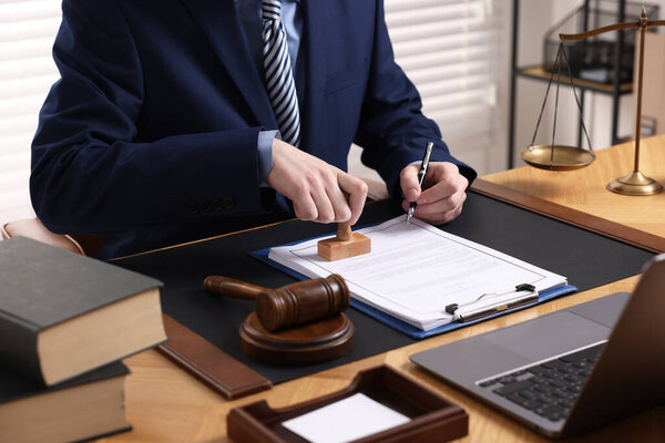 Notary with pen stamping document at wooden table in office, closeup