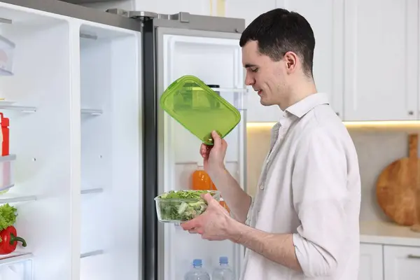 Happy man holding container with vegetables near refrigerator in kitchen
