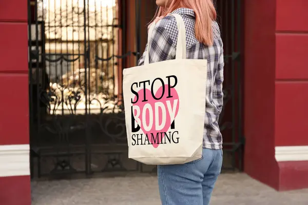 Woman holding bag with words Stop Body Shaming near building outdoors, closeup