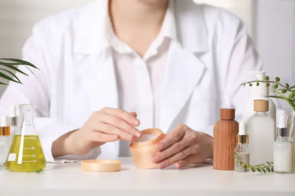 Dermatologist with jar testing cosmetic product at white table indoors, selective focus