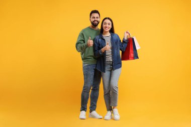 Alışveriş torbalı mutlu çift turuncu arka planda başparmak gösteriyor.