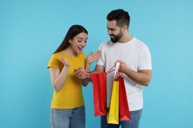 Bir adam açık mavi arka planda heyecanlı kız arkadaşına alışveriş torbası gösteriyor.
