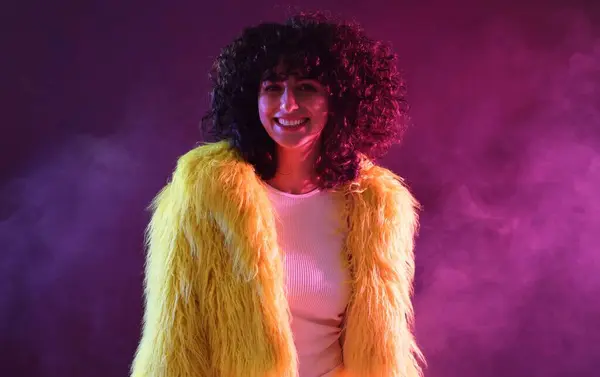 ネオンライトと煙の色の背景に黄色い毛皮のコートの美しい若い女性 — ストック写真