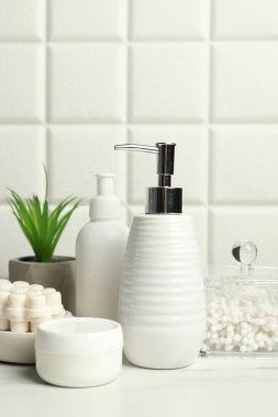 Duvarın yanındaki beyaz masada farklı banyo aksesuarları ve kişisel bakım ürünleri var.