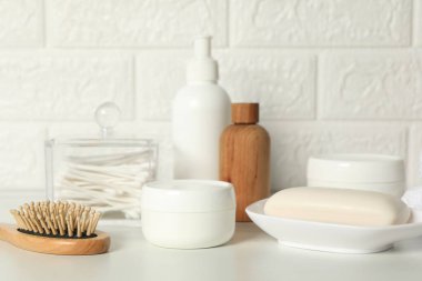 Farklı banyo aksesuarları ve kişisel bakım ürünleri tuğla duvarın yanındaki beyaz masada.