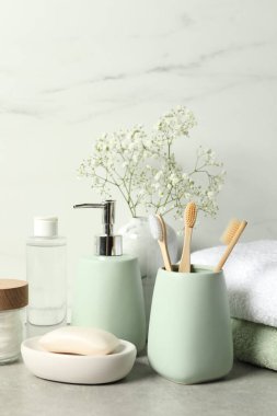 Farklı banyo aksesuarları, kişisel bakım ürünleri ve vazodaki çingene çiçekleri beyaz mermer duvarın yanındaki gri masada.