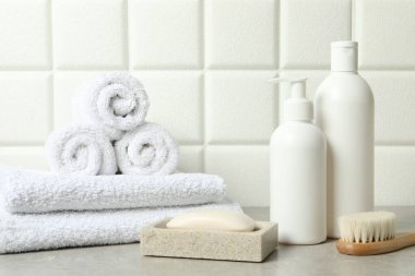 Farklı banyo aksesuarları ve kişisel bakım ürünleri beyaz fayanslı duvarın yanındaki gri masada.