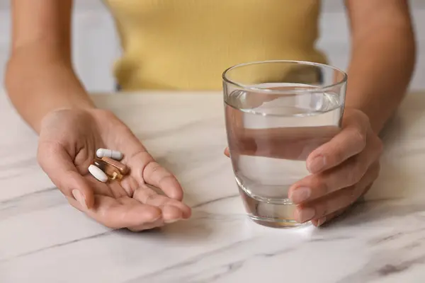 Woman with vitamin pills and glass of water at white marble table, closeup