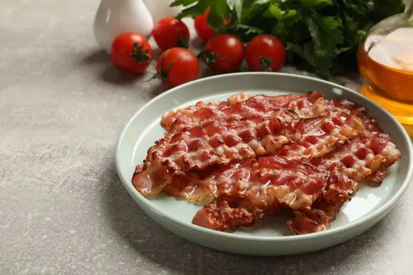 Plate with fried bacon slices, tomatoes and parsley on grey textured table, closeup