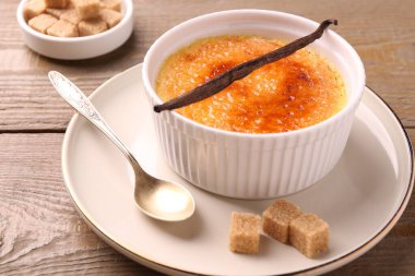 Lezzetli creme brulee kasede, vanilya kapsülü, kaşık ve şeker küpleri ahşap masada, yakın plan