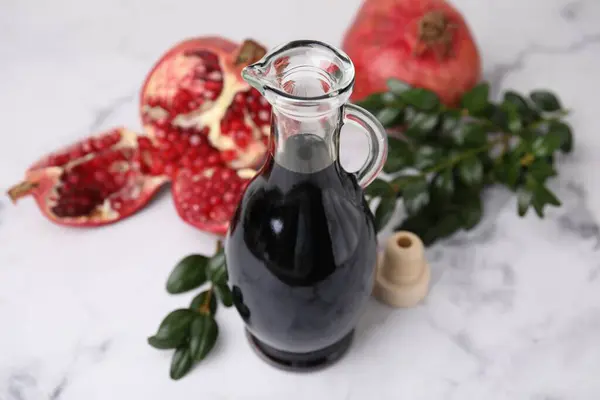 Tasty pomegranate sauce in bottle, fruits and branches on white marble table, closeup
