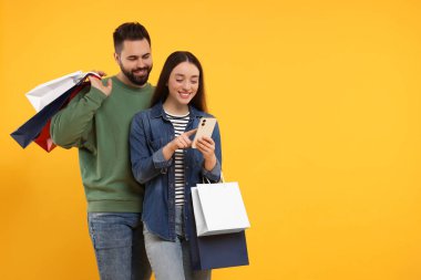 Alışveriş torbaları ve turuncu arka planda akıllı telefonları olan mutlu bir çift. Metin için boşluk