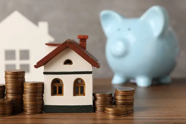 House models, piggy bank and stacked coins on wooden table against gray background, selective focus