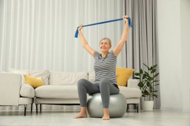 Kıdemli kadın evde spor salonunda elastik direniş bandıyla egzersiz yapıyor.